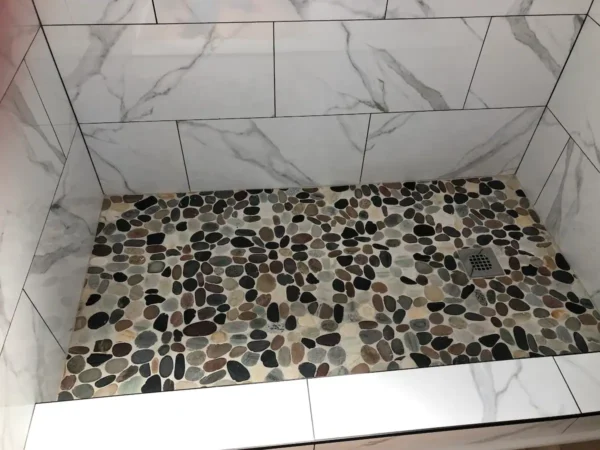 A shower with white tile and black, brown and gray stones.